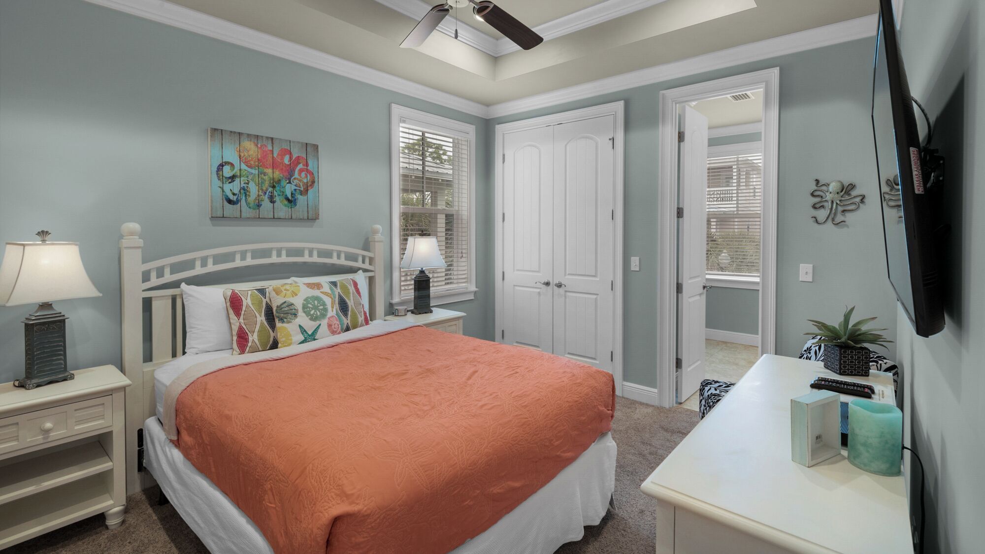 A bedroom with a bed, nightstands, a desk, a ceiling fan, and a door leading to another room.