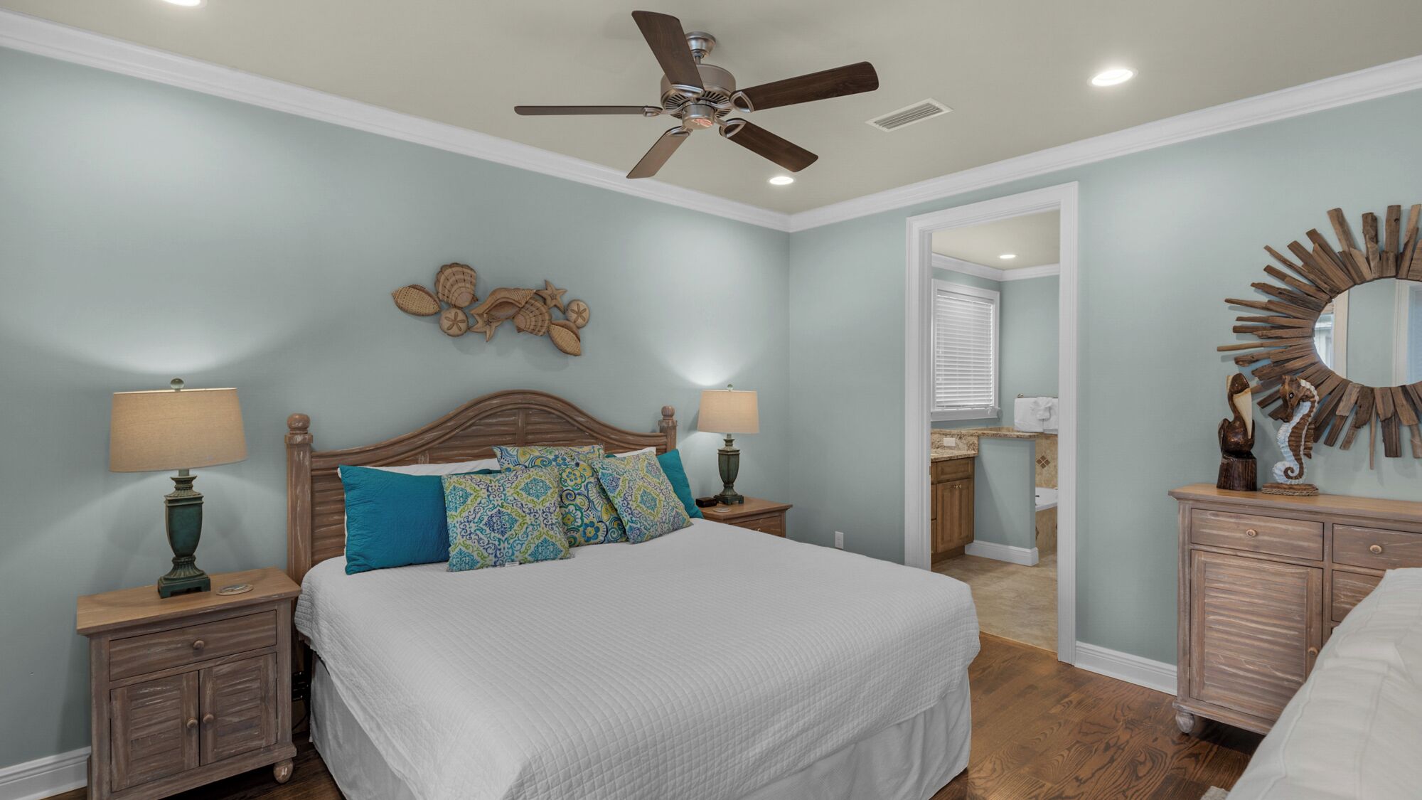 A bedroom with a bed, two nightstands, a ceiling fan, a wall-mounted mirror, and a bathroom visible through an open door.
