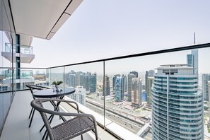 A modern balcony with a glass railing, a round table, and two wicker chairs. It overlooks a cityscape with high-rise buildings and a highway.