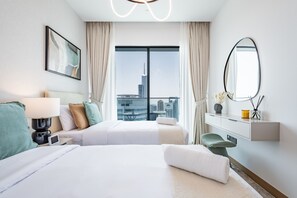 A hotel room with two beds, a large window with a city view, a bedside table with a lamp, a mirror, and a small desk with a chair.