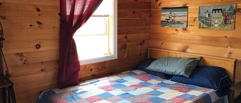 Habitació de cabana de fusta amb un llit, una finestra amb cortines vermelles i dos quadres a la paret.