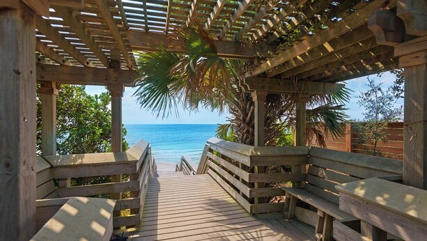 Une terrasse en bois qui mène à une plage, surmontée d’une pergola en bois.