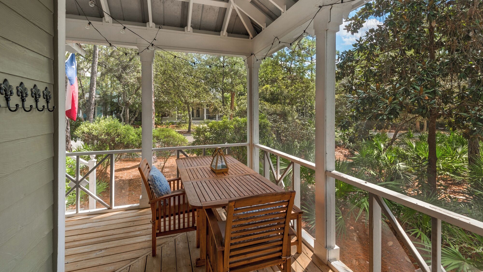 A covered outdoor area with a wooden table and chairs, surrounded by a white railing and lush greenery.