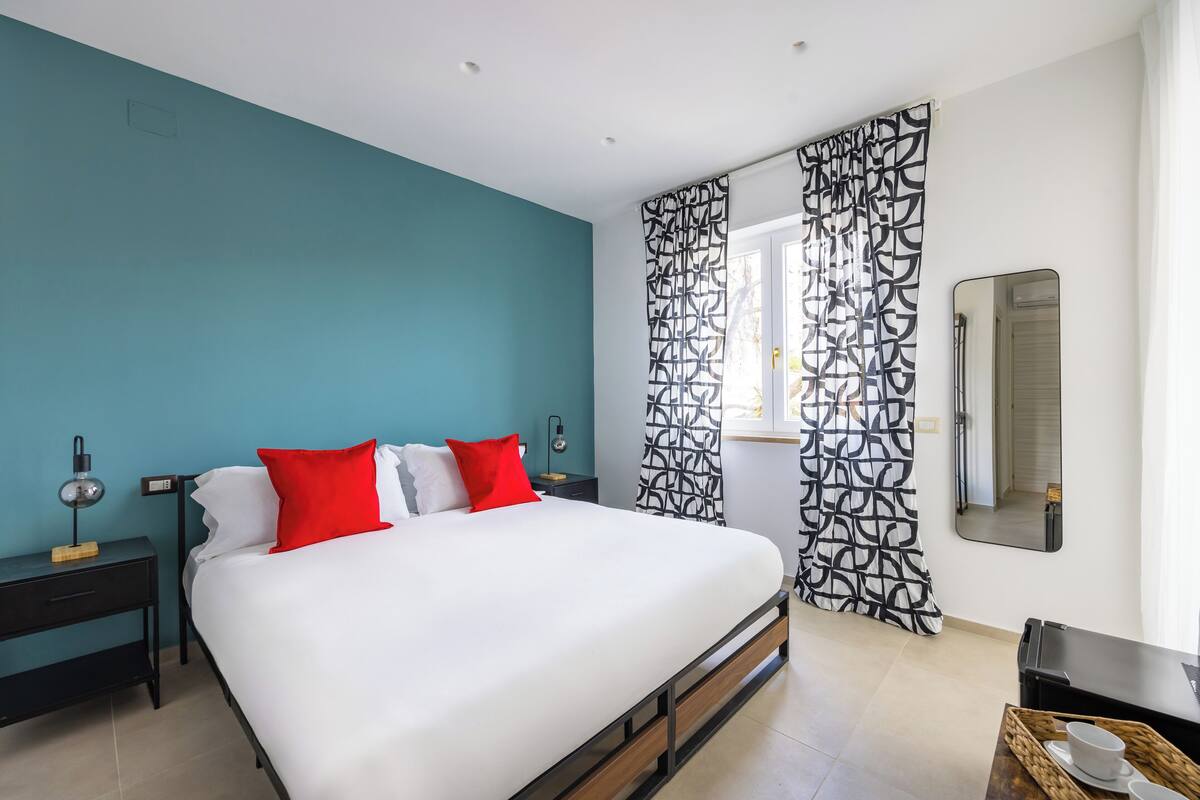 A modern bedroom with a teal accent wall, a large bed with white and red bedding, a black and white patterned curtain, and a bedside table with a lamp.