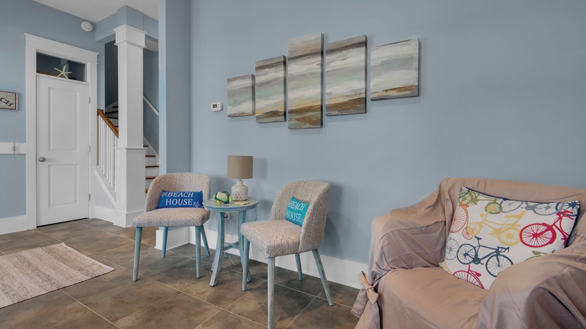 Una sala de estar con una escalera, dos sillones estampados con almohadas que dicen 'BEACH HOUSE', una mesa redonda con una lámpara y una pared decorada con cuatro cuadros abstractos.