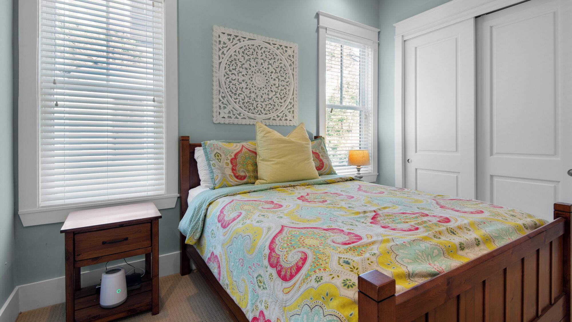 A bedroom with a wooden bed, a nightstand, a decorative wall piece, and a window with blinds.