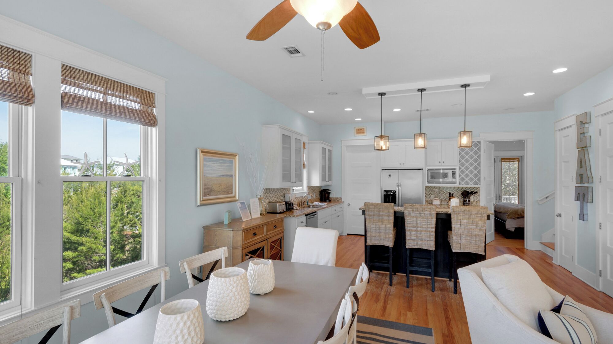 A modern kitchen with white cabinets, a dining area with a gray table, and a ceiling fan.