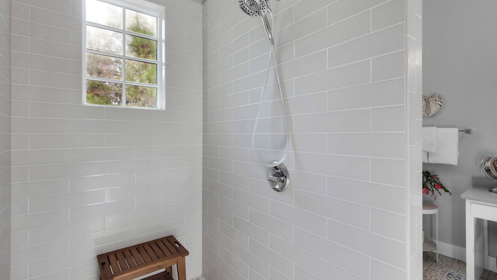 A bathroom with white tiled walls, a shower with a handheld showerhead, a small wooden bench, and a window with a view of trees.