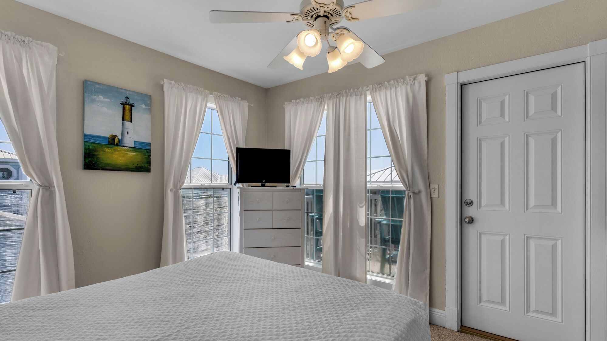 A bedroom with a bed, a TV, a ceiling fan, and a painting of a lighthouse.
