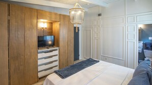 Ett sovrum med en säng, en tv-apparat placerad på ett träskåp, en kristallkrona och ett badrum som syns genom en öppen dörr.