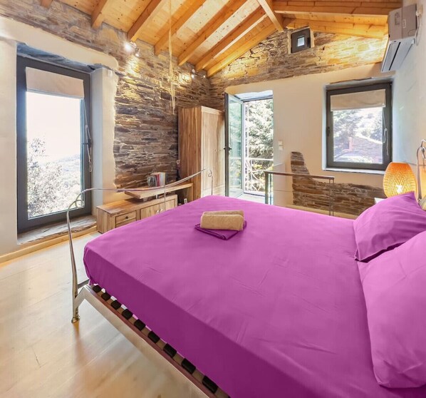 Une chambre avec un grand lit, des murs en pierre, des poutres en bois et de grandes fenêtres.