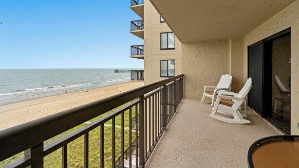 Un balcon avec vue sur la plage et l’océan, comprenant une balustrade, une table et deux fauteuils à bascule.
