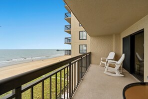 A balcony with a view of the beach and ocean, featuring a railing, a table, and two rocking chairs.