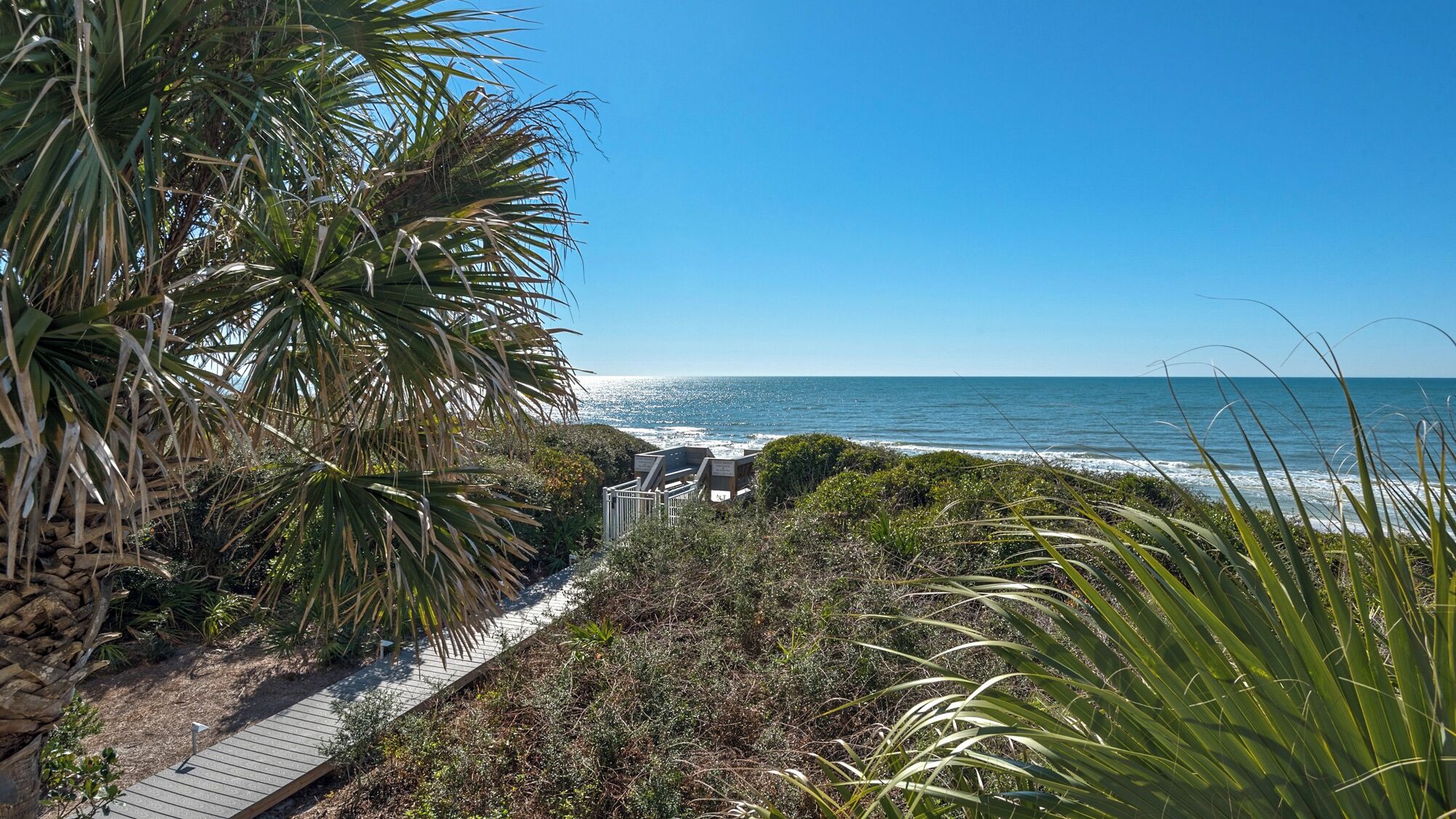 A coastal pathway with a wooden boardwalk, leading to a beach with ocean views.