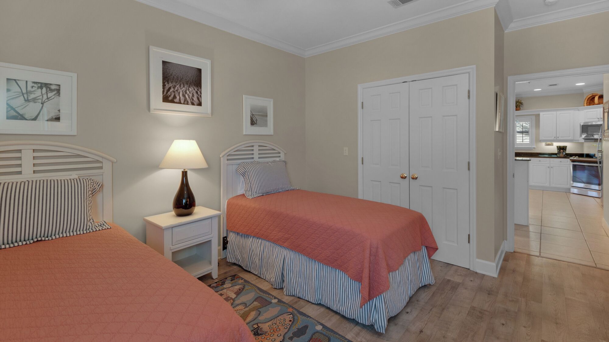 A bedroom with two beds, a nightstand with a lamp, a door, and a view into a kitchen.