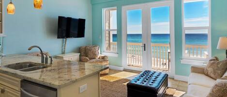 A living room with a large window overlooking the ocean, a kitchen with a sink and a dishwasher, and a seating area with a sofa and ottoman.