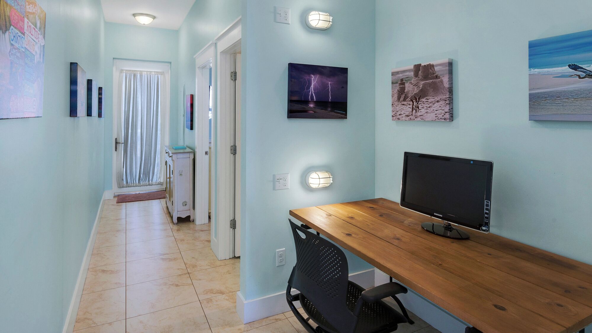 A hallway with a wooden desk, a computer, and a chair. There are wall-mounted lights and framed pictures.
