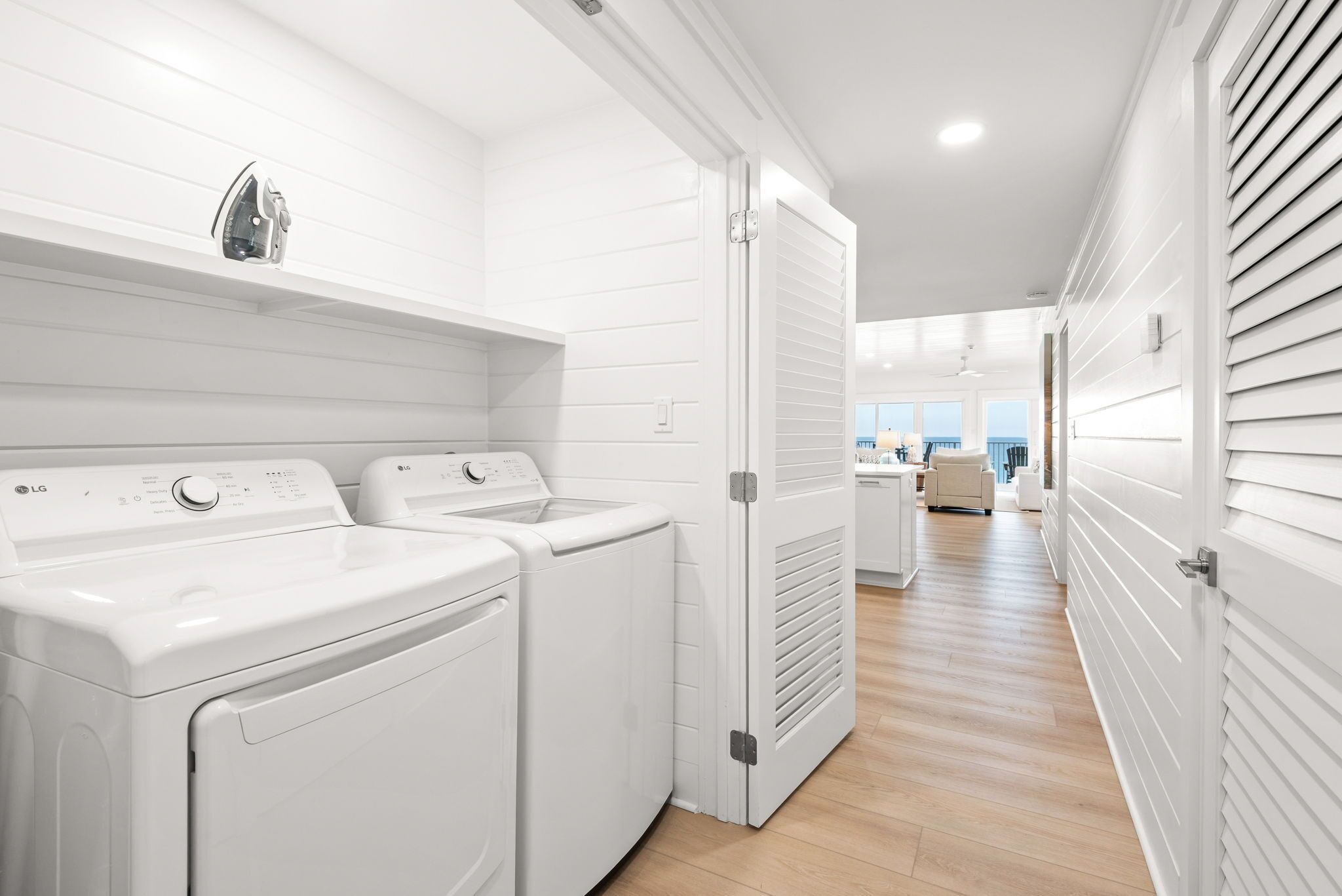 Une buanderie équipée de deux lave-linge et deux sèche-linge blancs, d’un fer à repasser fixé au plafond, et d’un couloir menant à l’espace de vie.