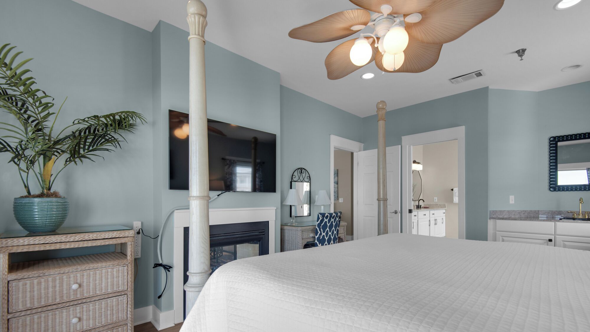 A bedroom with a large bed, a ceiling fan, a potted plant, a fireplace, and a bathroom with a mirror and sink.