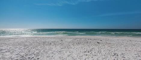 A sandy beach with footprints leading to the water, a clear sky, and a distant jet trail.