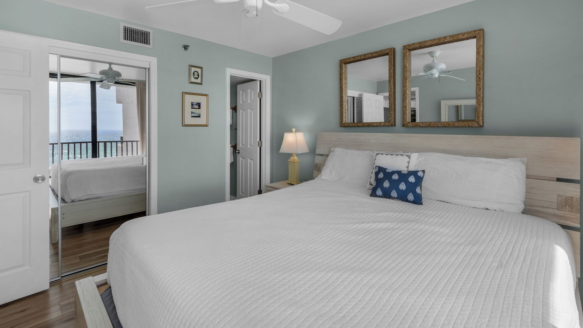 A bedroom with a bed, two wall mirrors, a ceiling fan, and a view of the ocean through an open door.