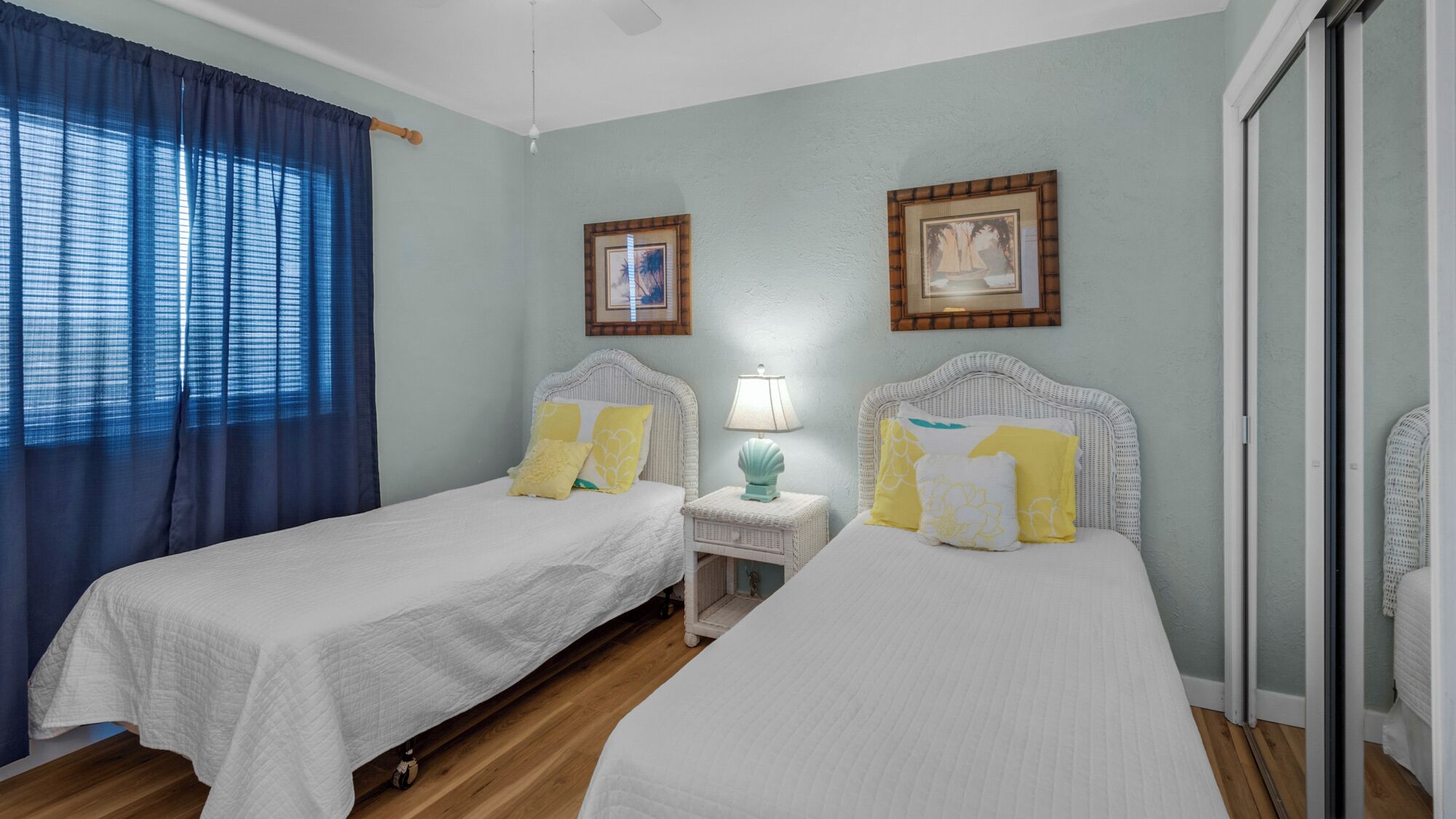 A hotel room with two beds, a nightstand with a lamp, and a large window with blue curtains.