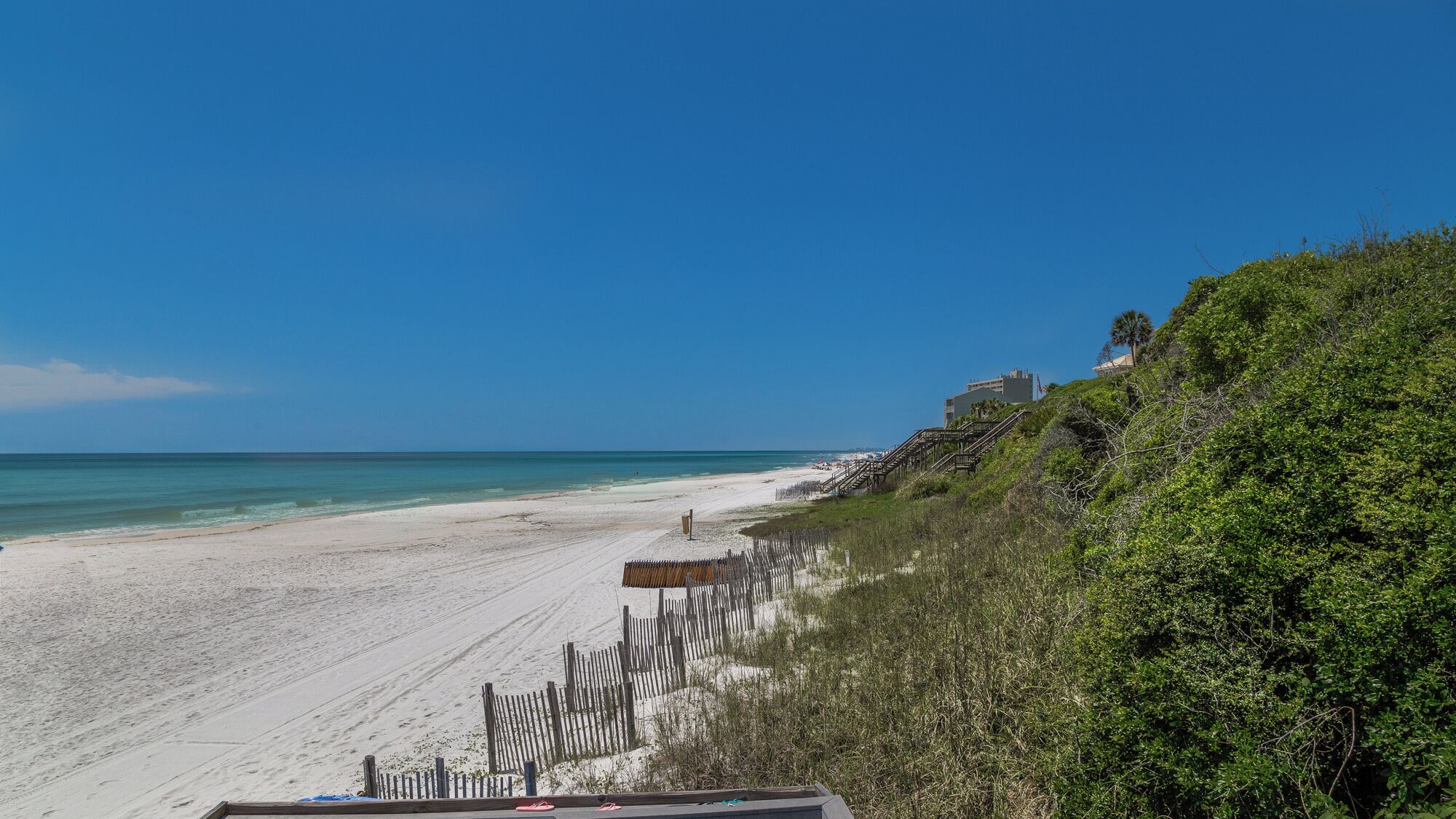 A beach with a wooden fence, a clear blue sky, and a building on a hill.