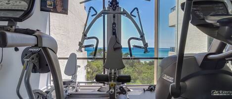 A home gym with various exercise equipment, including a treadmill, a stationary bike, and a weight machine, with a view of the ocean through large windows.