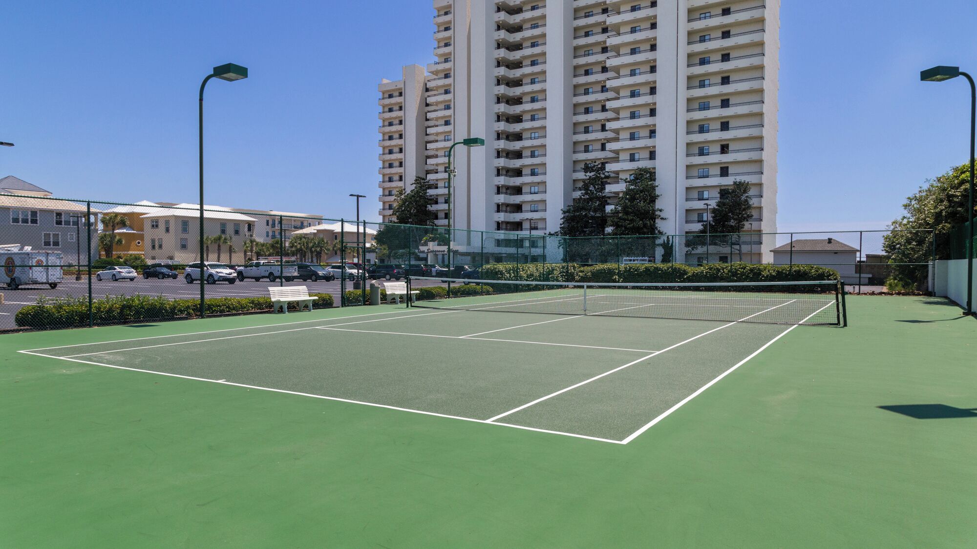 Un campo da tennis all'aperto con rete, circondato da una recinzione e adiacente a un'area di parcheggio con diverse auto.