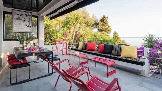 An outdoor seating area with red chairs and a bench, surrounded by greenery and a view of the sea.