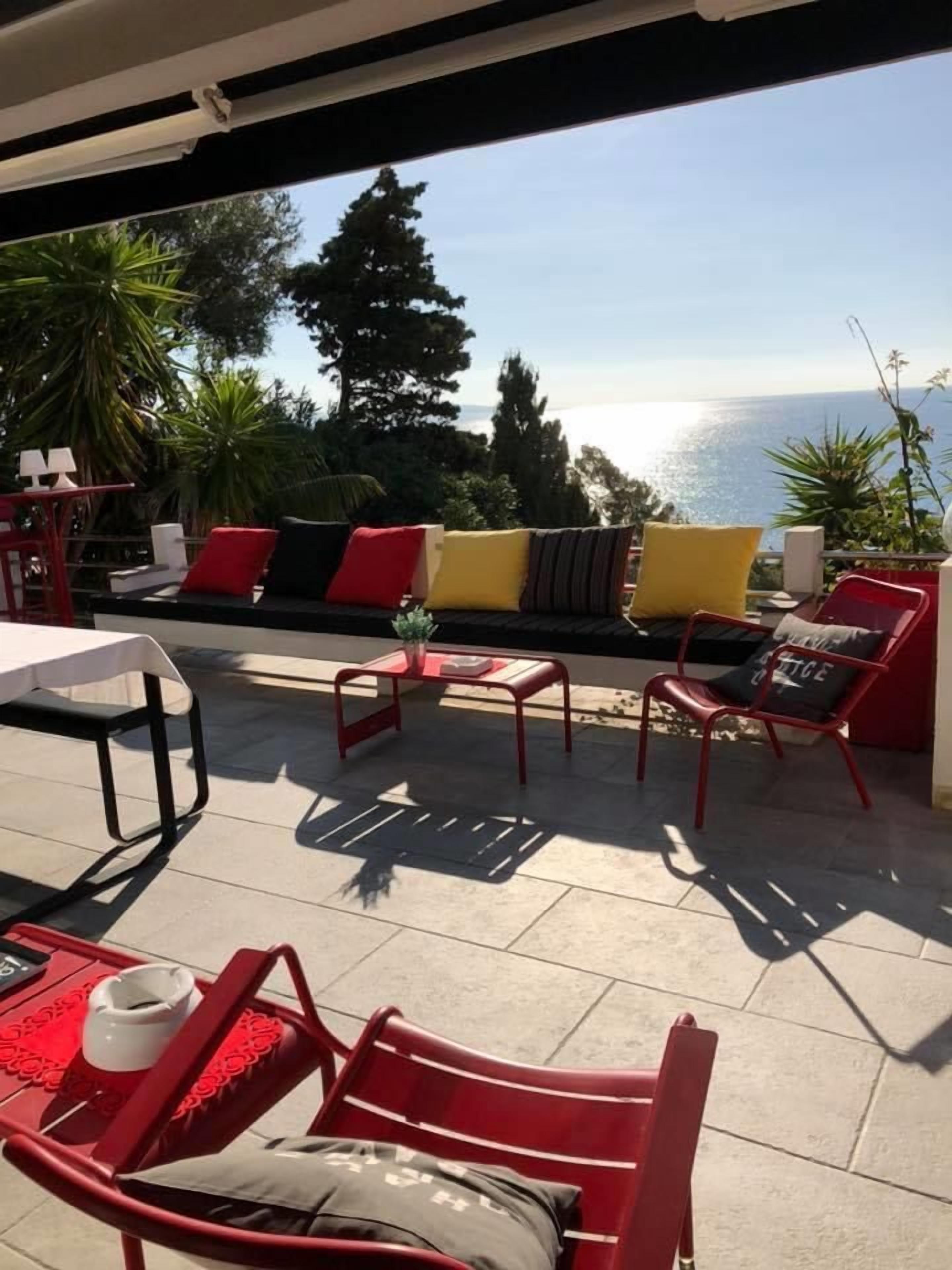 An outdoor patio with red furniture, a white table, and a view of the sea.