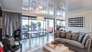 A modern living room with a sofa, coffee table, and chairs. There is a dining area with a table and chairs. A large window provides natural light, and a wall-mounted shelf displays decorative items.