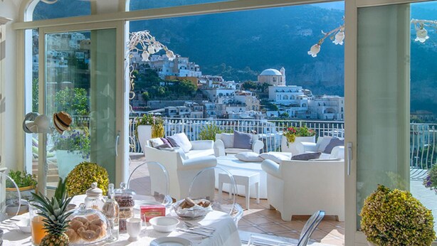 Zona pranzo con tavolo apparecchiato per quattro, affacciata su una località costiera con edifici bianchi e montagne sullo sfondo.