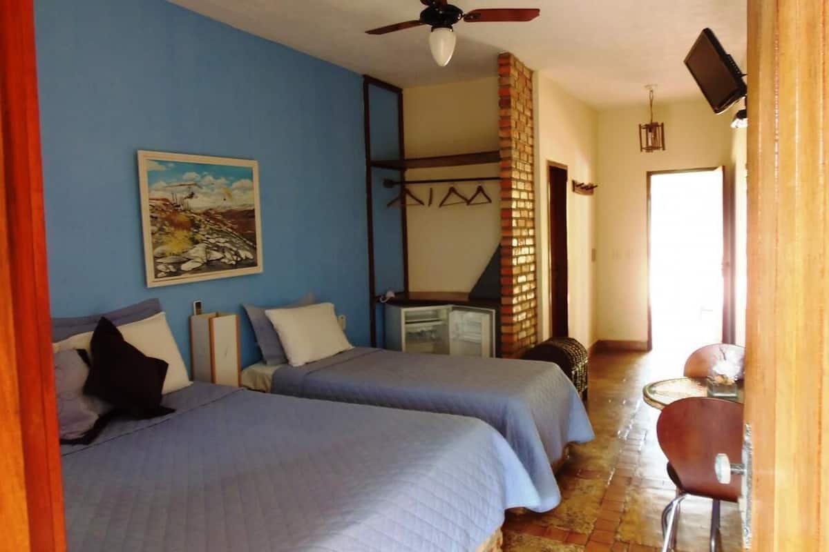 Una habitación con dos camas, un ventilador de techo, un cuadro en la pared, una mesita y una silla.