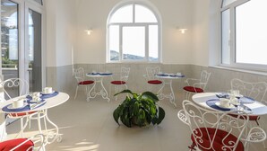 Un espace repas baigné de lumière naturelle, avec des tables et des chaises en métal blanc, une plante en pot au centre et de grandes fenêtres en arc de cercle offrant une vue sur le paysage.