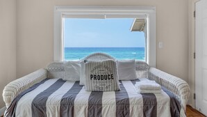 Une chambre avec une grande fenêtre donnant sur l’océan, un lit avec une literie à rayures et un oreiller orné de la mention : « PRODUITS LOCAUX FRAIS, AGRICULTURE BIOLOGIQUE, DEPUIS 1871 ».