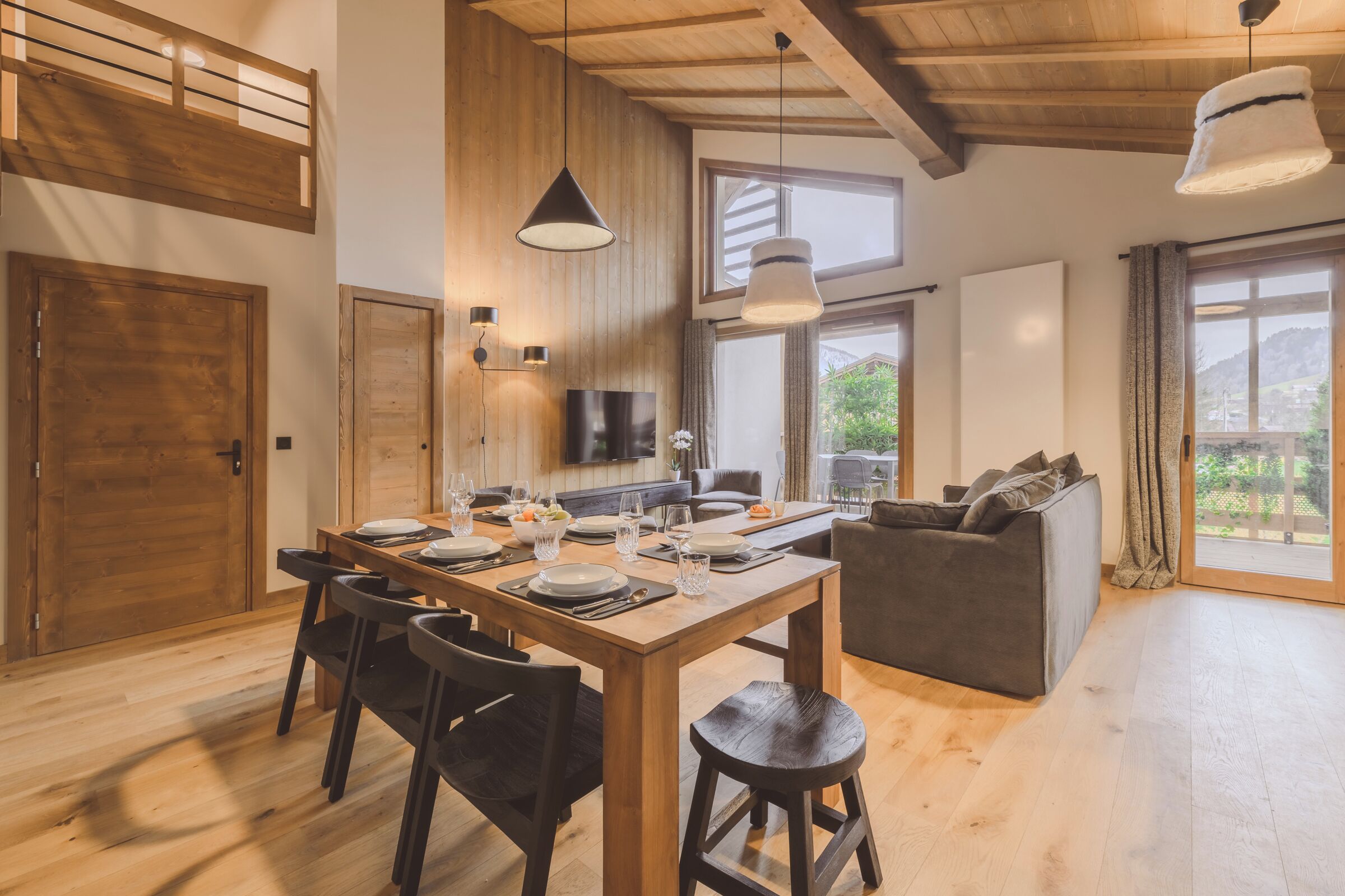 A spacious living area with a wooden dining table set for four, a gray sofa, and a TV mounted on the wall.