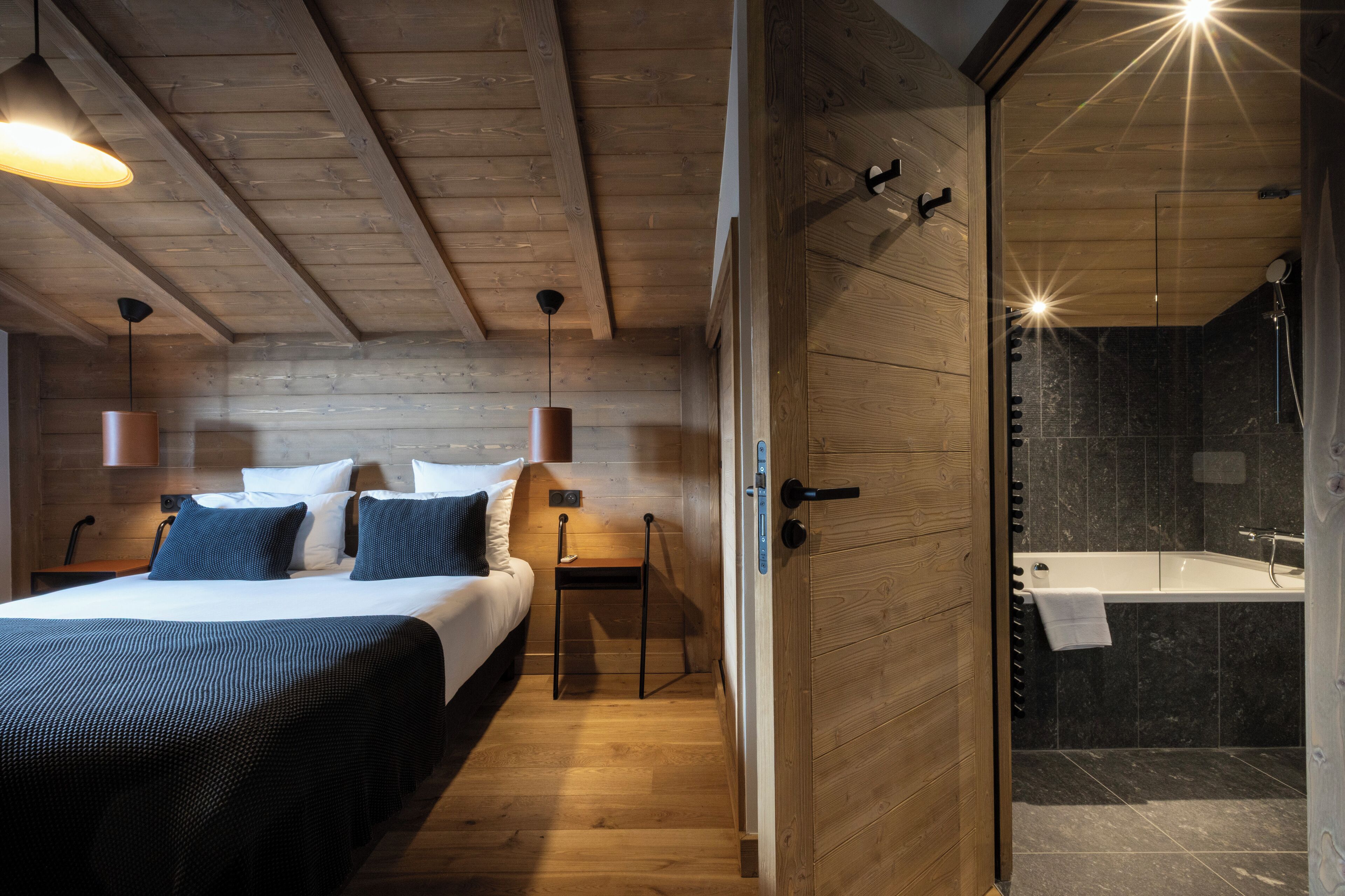 A wooden bedroom with a large bed, bedside tables, and a nightstand. There is a wooden door leading to a bathroom with a bathtub and a shower.