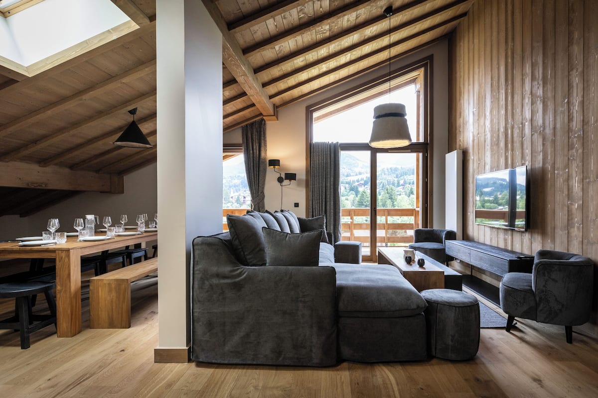 A modern living room with a sectional sofa, wooden dining table, and a large window offering a view of the outdoors.