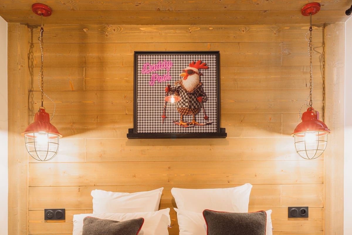 A neatly made bed with white and grey pillows, a framed picture of a rooster, and two red pendant lights.