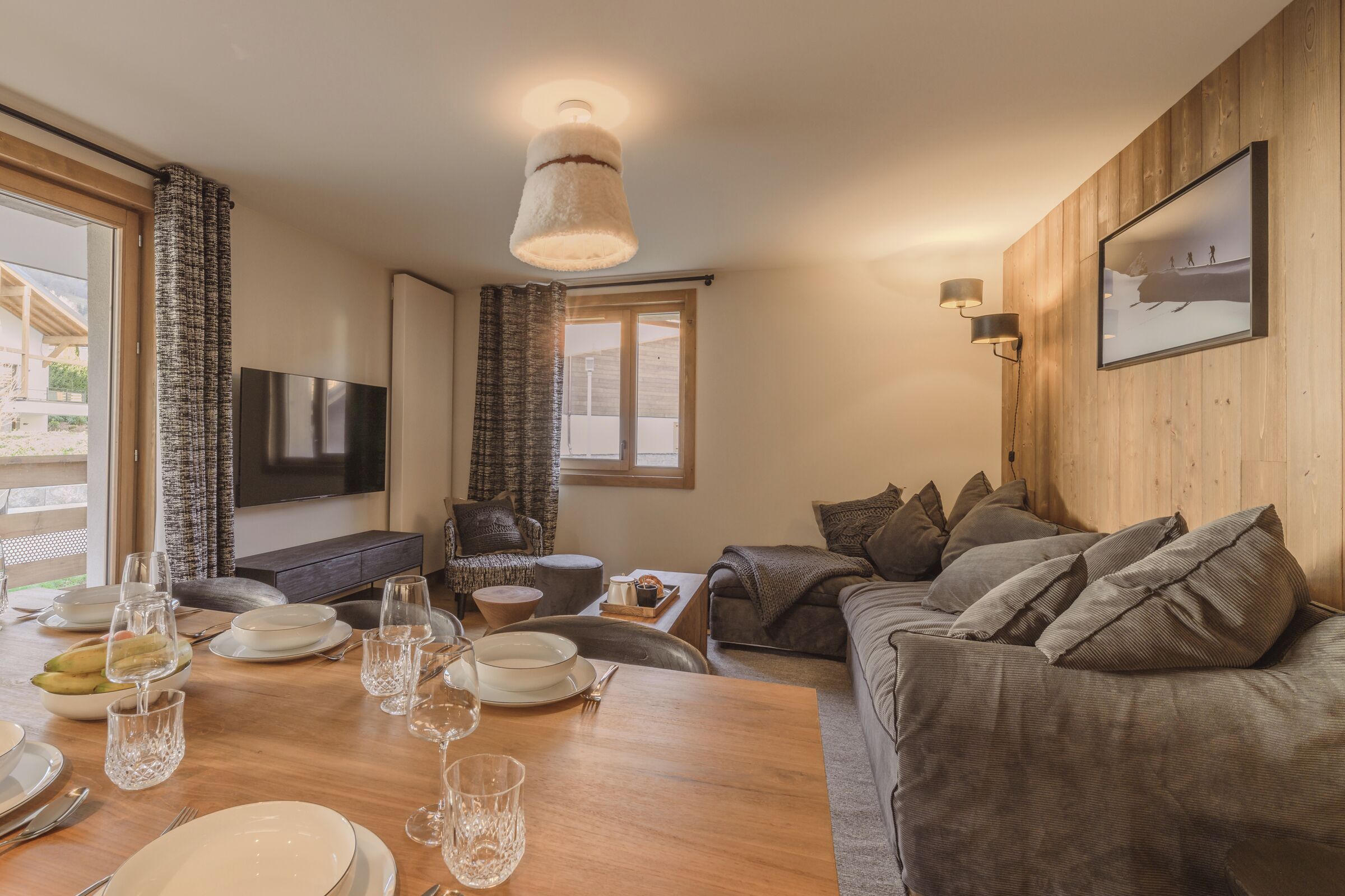 A modern living room with a wooden wall, a large flat-screen TV, a dining table set with plates and glasses, and a sofa with cushions.