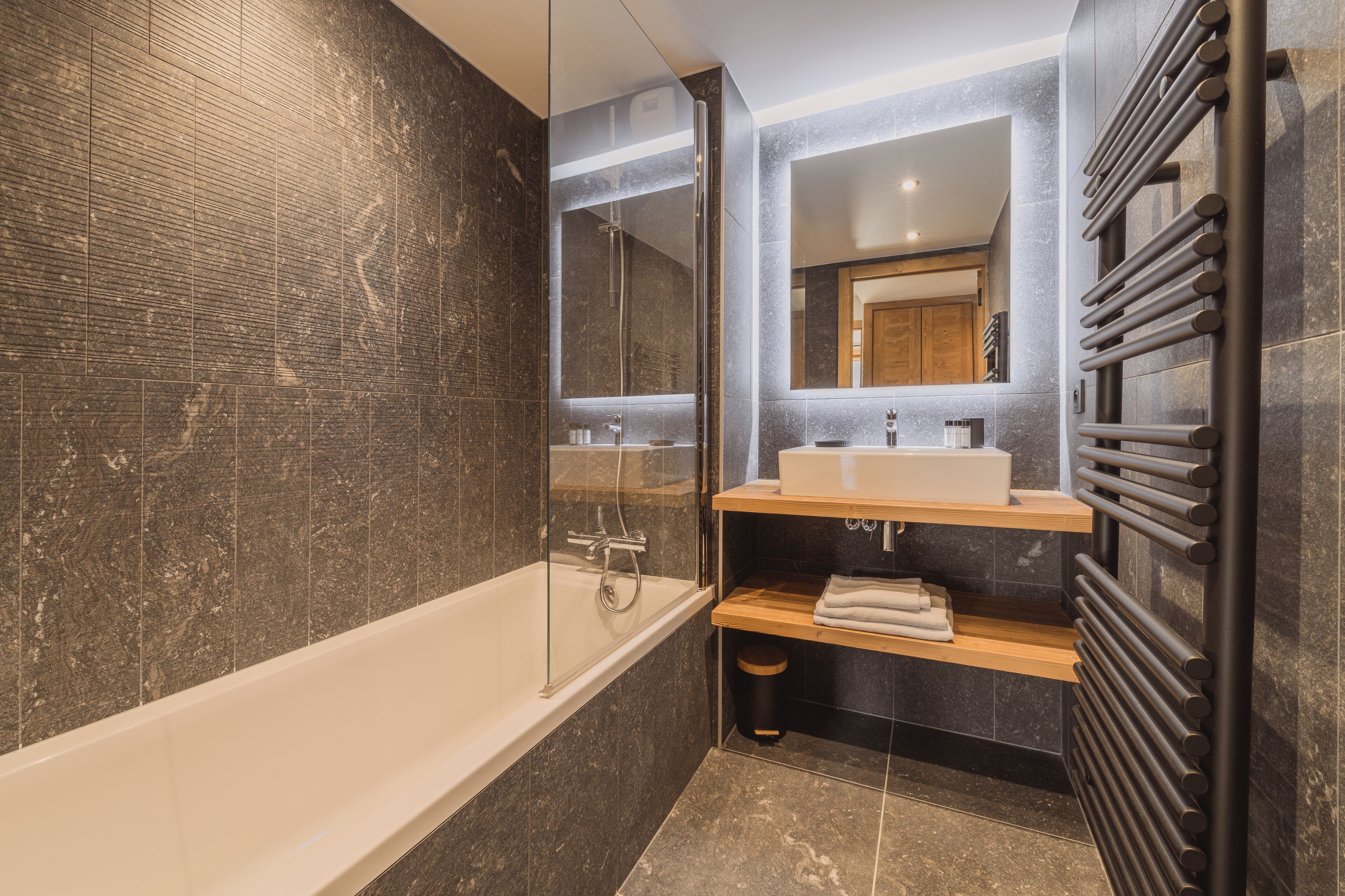 Un baño moderno con ducha de vidrio, bañera blanca, tocador de madera con espejo y toallero negro.