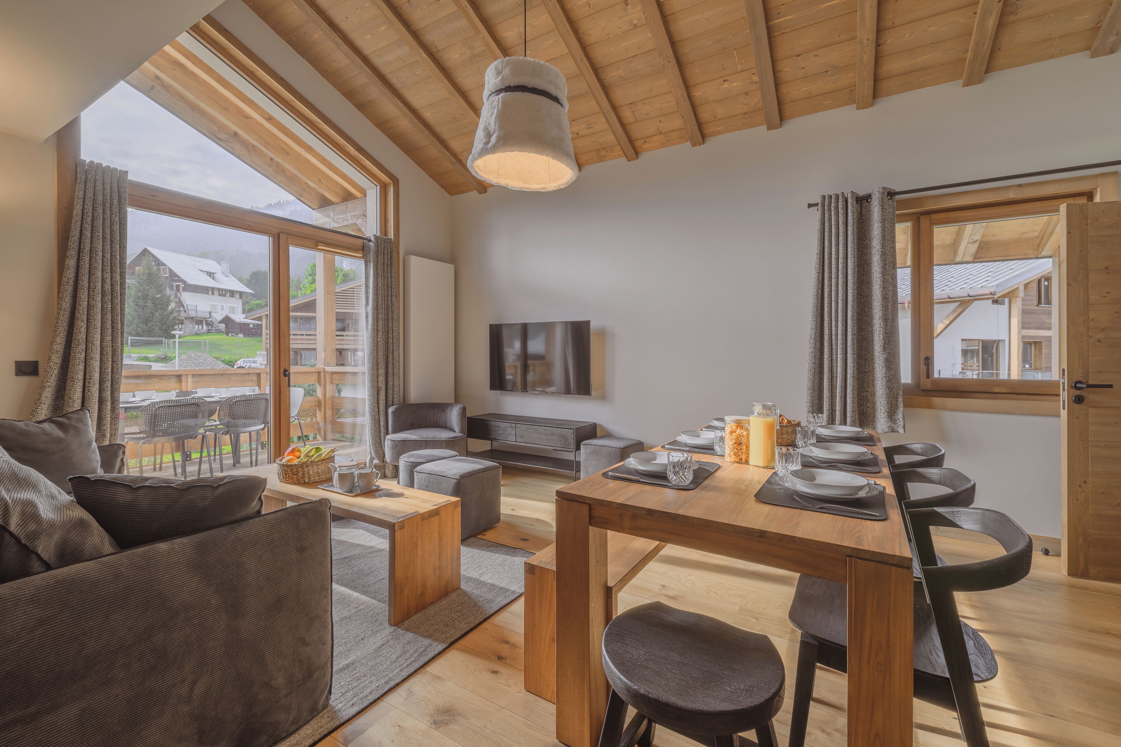 A modern living room with a wooden dining table, grey sofas, and a large window offering a view of a house and greenery.