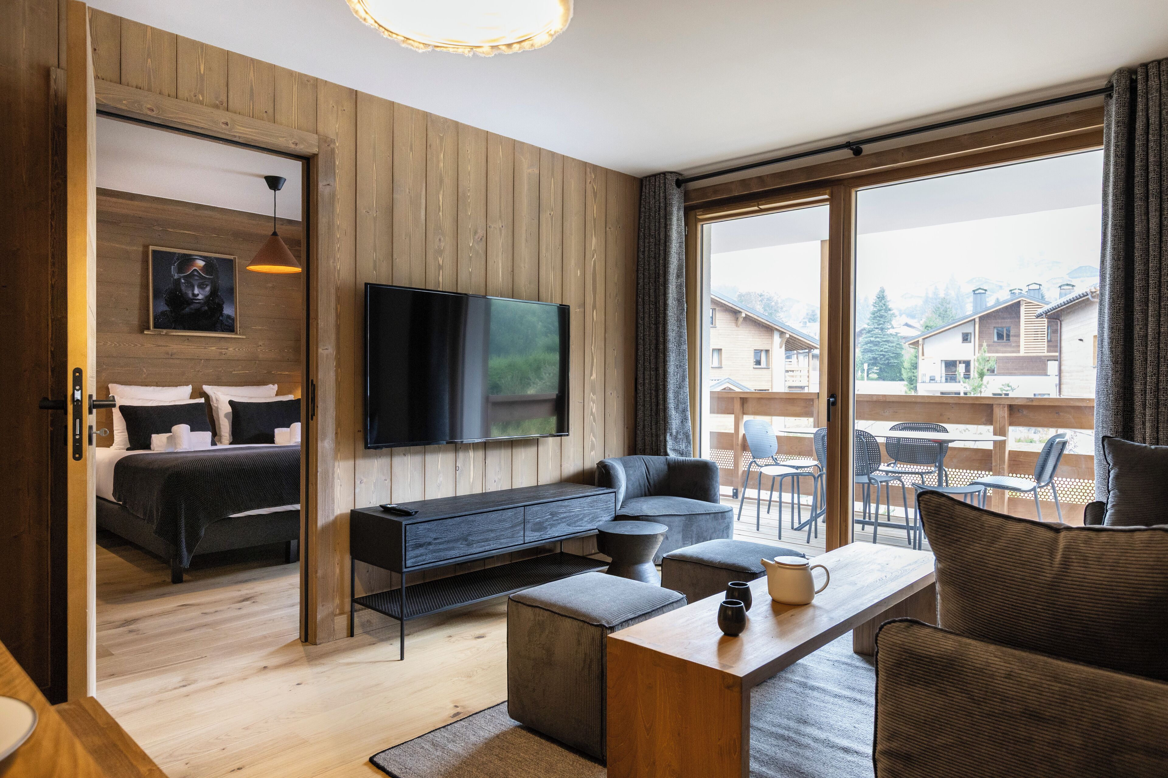Une chambre d’hôtel moderne avec un intérieur en bois, une grande fenêtre, un téléviseur à écran plat, un canapé, une table basse et un lit doté d’une tête de lit de couleur foncée.