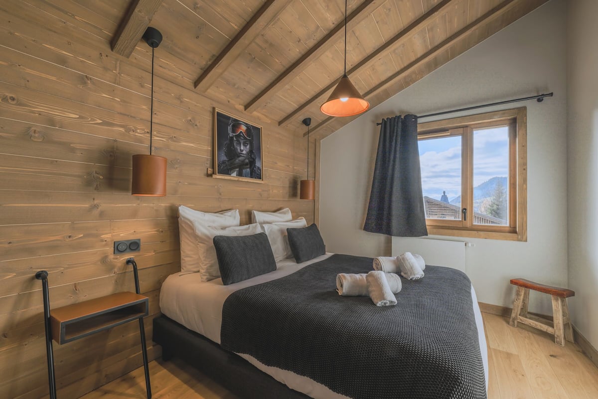 A wooden-paneled bedroom with a bed, a hanging pendant light, a window with a view, and a small wooden stool.
