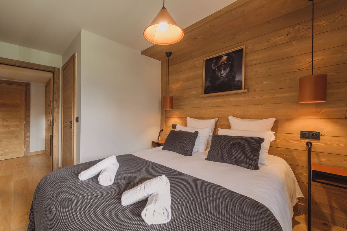 A neatly made bed with a grey and white patterned bedspread, a pair of rolled towels, and a framed picture on the wall above the bed.