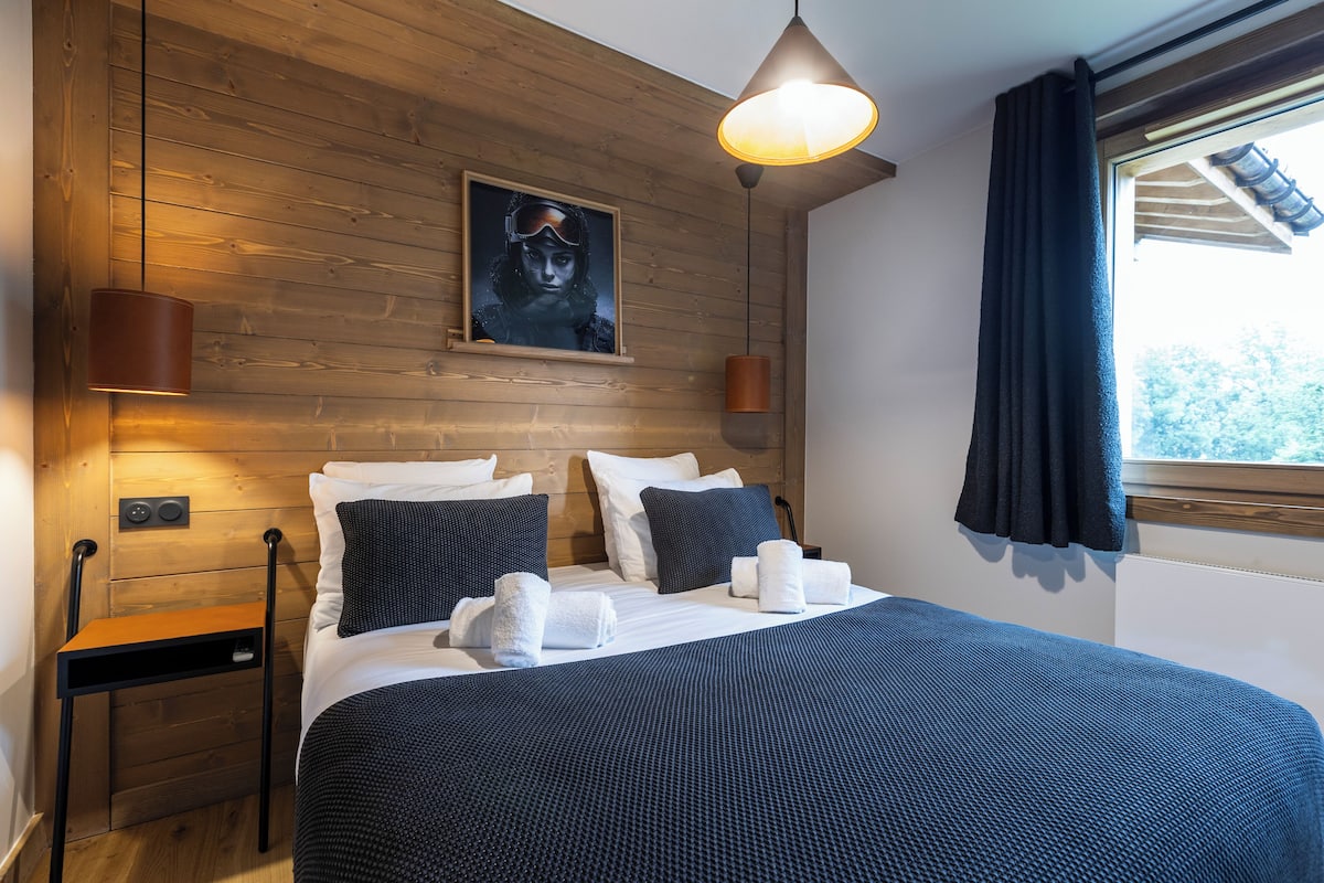 A neatly made bed with a dark blue bedspread, two pillows, and a folded towel. A wooden headboard and a framed picture are visible on the wall.