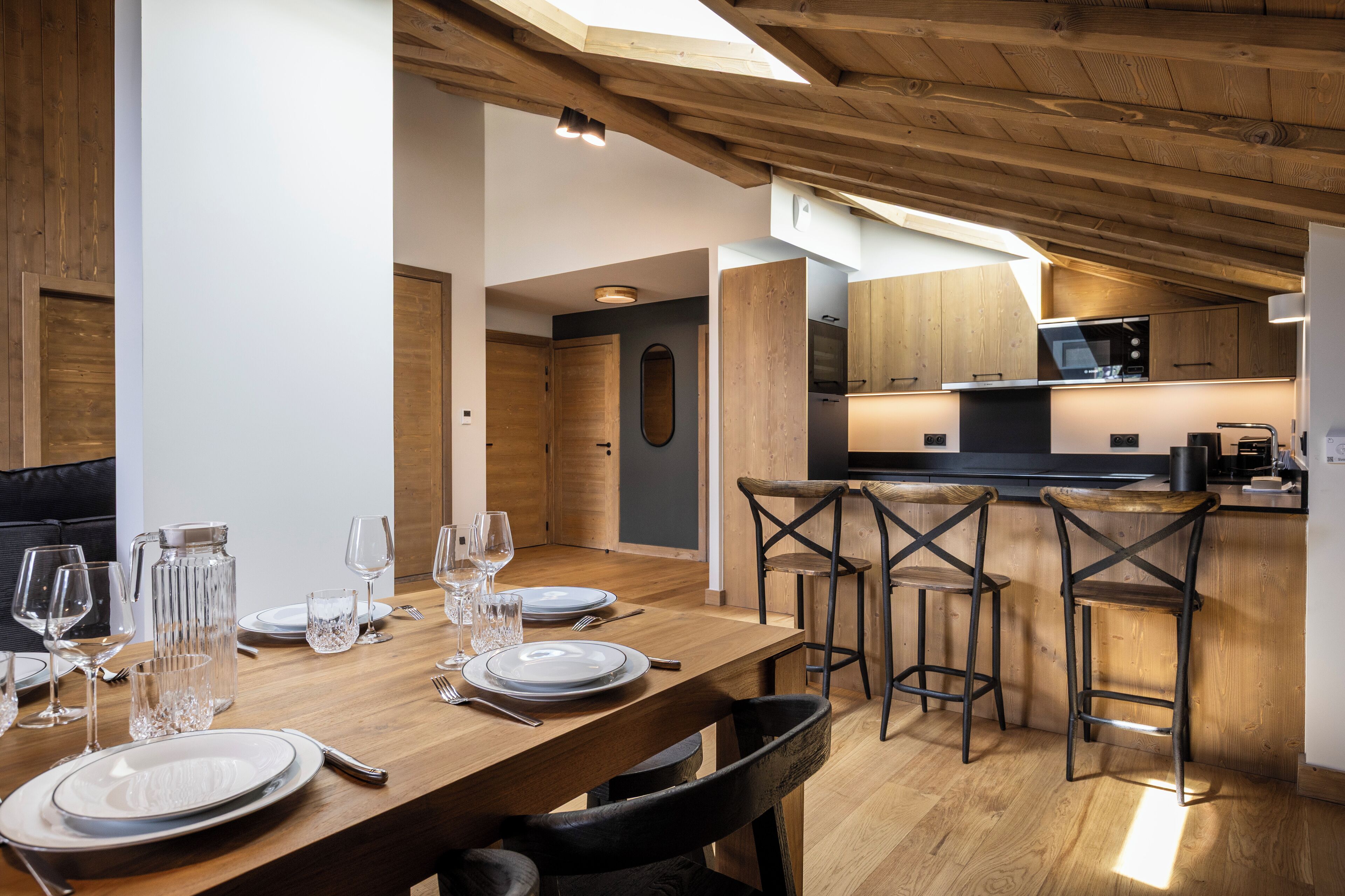 A dining area with a wooden table set for six, a kitchen in the background, and a high ceiling with exposed beams.