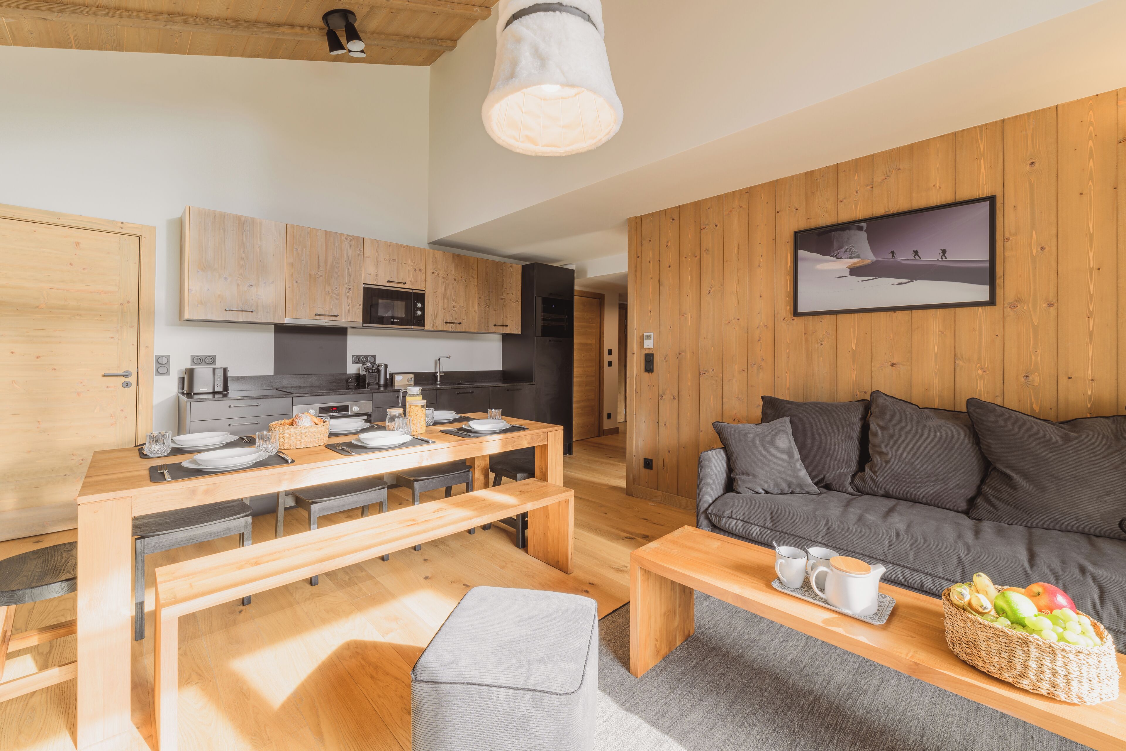 A modern kitchen with wooden cabinets, a dining area with a bench, and a living room with a sofa and a painting on the wall.