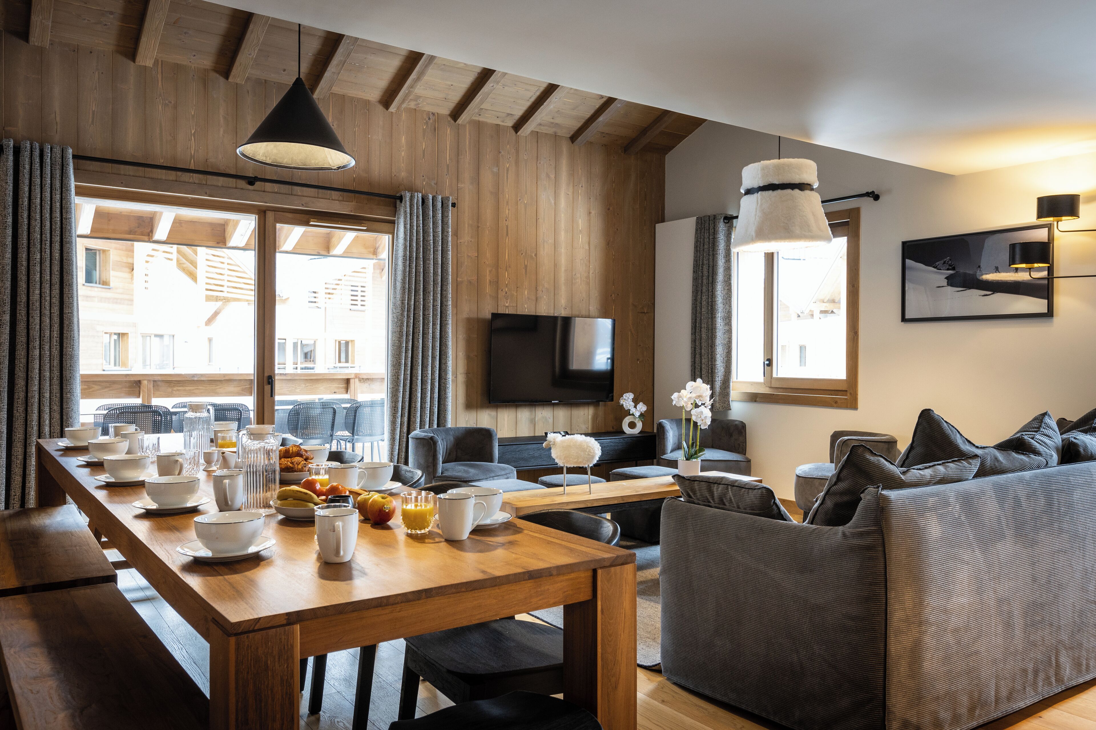 A modern living room with a wooden dining table set for breakfast, a grey sofa, and a flat-screen TV mounted on the wall.